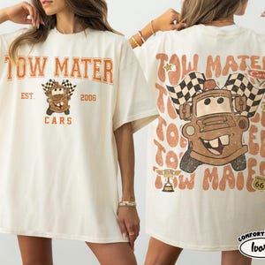 May include: Ivory-colored t-shirt featuring a graphic of Tow Mater from the Cars movie. The front of the shirt displays "Tow Mater Cars" in orange lettering, with "Est. 2006" and a cartoon image of Mater. The back has a larger Mater graphic and text.