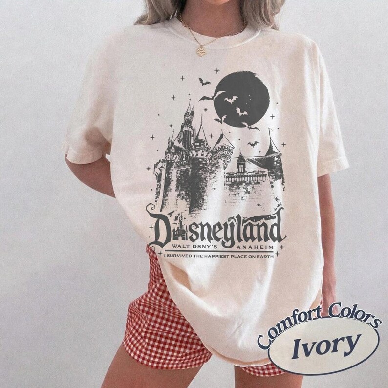 K&ouml;nnte beinhalten: Elfenbeinfarbenes T-Shirt mit einer Grafik eines Schlosses, Flederm&auml;usen und einem Vollmond. Unter dem Schlossbild ist der Text "Disneyland Anaheim" mit dem Satz "I survived the happiest place on earth" aufgedruckt.