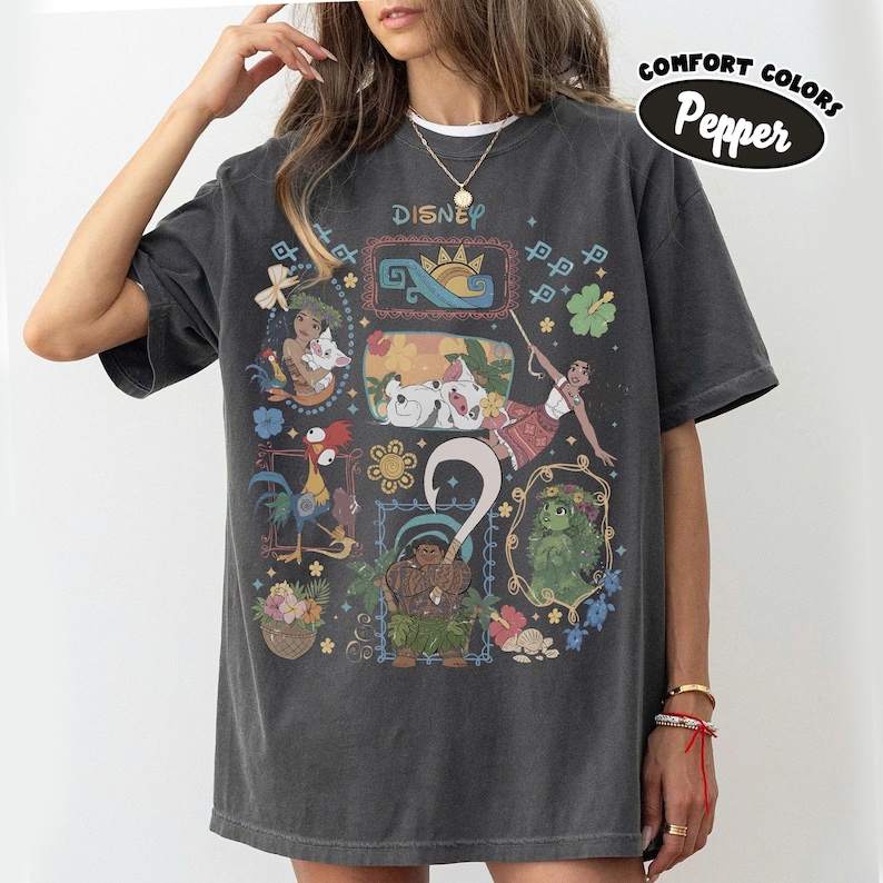 May include: Dark gray t-shirt featuring a colorful graphic of Disney's Moana characters. The design includes illustrations of Moana, Maui, Hei Hei, and Pua, framed with floral and geometric elements. The word "Disney" is printed at the top.