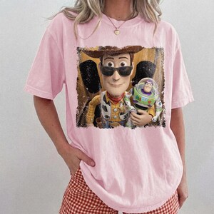 K&ouml;nnte beinhalten: Ein hellrosa T-Shirt mit einem Grafikdruck eines Cowboy-Charakters, der eine schwarze Sonnenbrille tr&auml;gt und eine Weltraum-Actionfigur h&auml;lt. Das Motiv hat einen abgenutzten rechteckigen Rand und zeigt die Figuren in Gelb, Braun, Gr&uuml;n und Violett.