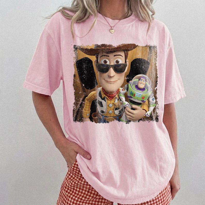 K&ouml;nnte beinhalten: Ein hellrosa T-Shirt mit einem Grafikdruck eines Cowboy-Charakters, der eine schwarze Sonnenbrille tr&auml;gt und eine Weltraum-Actionfigur h&auml;lt. Das Motiv hat einen abgenutzten rechteckigen Rand und zeigt die Figuren in Gelb, Braun, Gr&uuml;n und Violett.