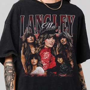 Puede incluir: Una camiseta negra de manga corta con un estampado gráfico que incluye el texto LANGLEY y Ella en una fuente estilizada. El diseño presenta cinco retratos fotográficos de la misma persona con cabello largo castaño y flequillo, sobre un fondo rojo.