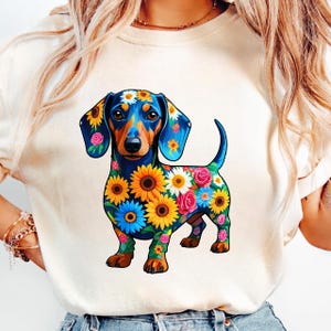 Könnte beinhalten: Ein cremefarbenes T-Shirt mit einer farbenfrohen Illustration eines Dackelhundes, der mit Sonnenblumen, Gänseblümchen und Rosen geschmückt ist. Der Hund hat einen blau-orangen Körper mit floralen Akzenten, was ein lebendiges Design ergibt. Das T-Shirt ist ein Freizeitartikel.
