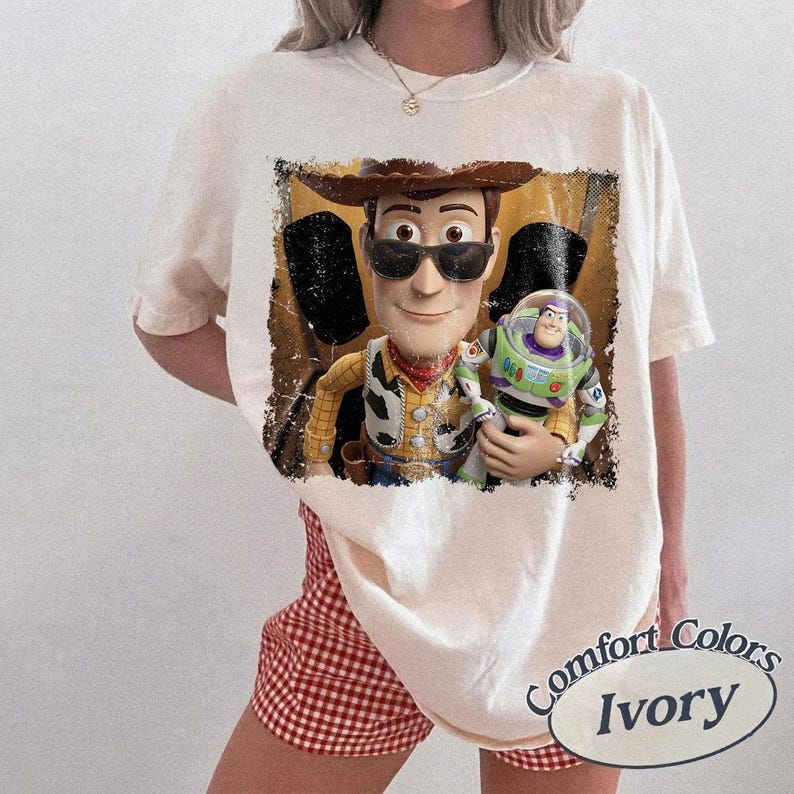 K&ouml;nnte beinhalten: Ein kurz&auml;rmeliges T-Shirt in Elfenbein mit einem abgenutzten Grafikdruck der Figuren Woody und Buzz Lightyear. Woody tr&auml;gt eine dunkle Sonnenbrille und einen Cowboyhut. Die Grafik hat ein strukturiertes, abgenutztes Aussehen.