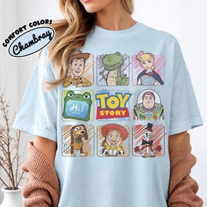 May include: Pale blue t-shirt displaying a grid of Toy Story characters, such as Woody, Rex, and Buzz Lightyear. The shirt also features the text "TOY STORY" and "Comfort Colors Chambray" in a circular design.