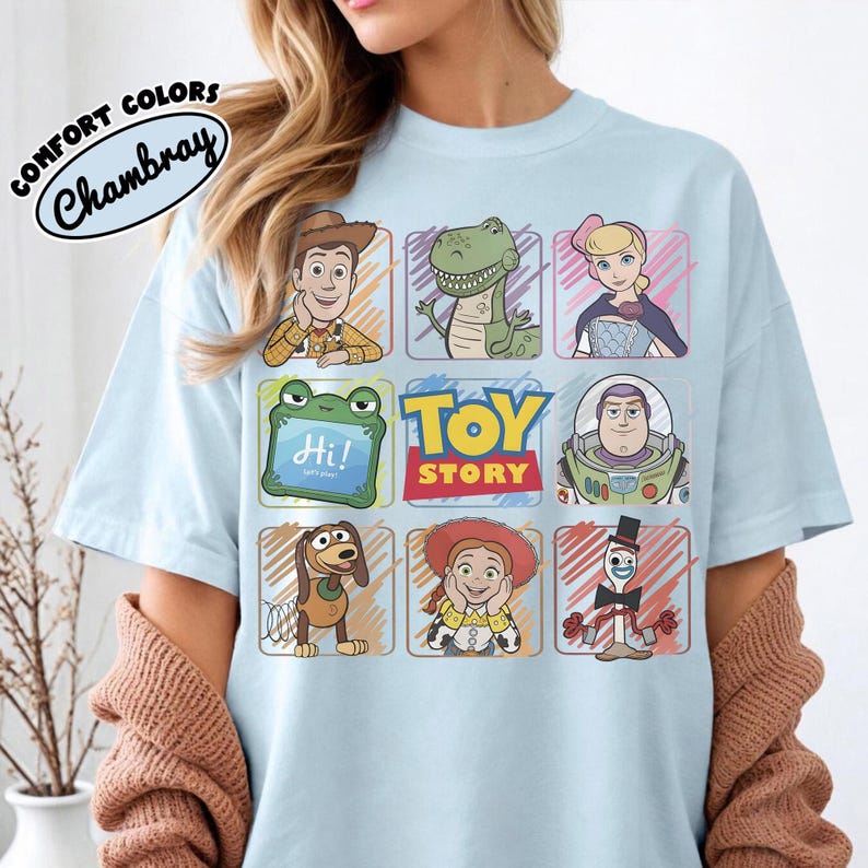 May include: Pale blue t-shirt displaying a grid of Toy Story characters, such as Woody, Rex, and Buzz Lightyear. The shirt also features the text "TOY STORY" and "Comfort Colors Chambray" in a circular design.