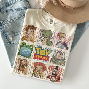 May include: Cream-coloured t-shirt with a colourful Toy Story graphic. The design includes characters such as Woody, Rex, and Buzz Lightyear. The shirt is displayed on light blue denim jeans and a tan hat.
