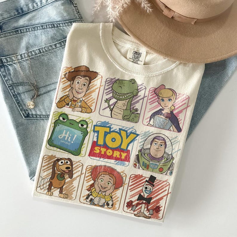 May include: Cream-coloured t-shirt with a colourful Toy Story graphic. The design includes characters such as Woody, Rex, and Buzz Lightyear. The shirt is displayed on light blue denim jeans and a tan hat.