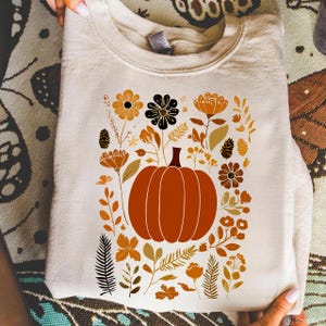 Könnte beinhalten: Cremefarbenes Sweatshirt mit einem großen orangefarbenen Kürbis mit braunem Stiel, umgeben von floralen und Blattillustrationen in Orange-, Braun- und Schwarztönen. Das Design ist auf der Vorderseite des Shirts zentriert.