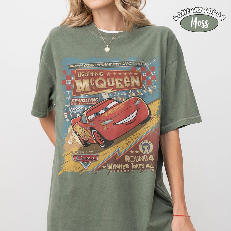 May include: A moss green short-sleeve t-shirt featuring a graphic print of the character Lightning McQueen from the movie Cars. The design includes the text Radiator Springs Saturday Night Special, Lightning McQueen, Re-Volting, and Round 4 Winner Takes All.