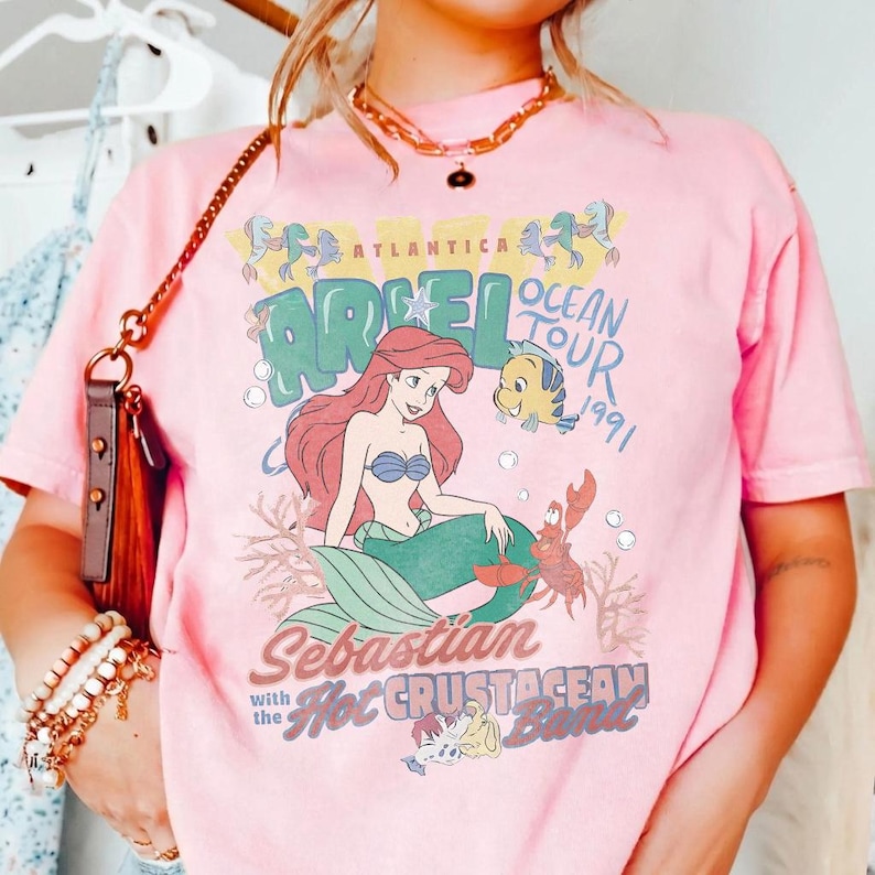 May include: A light pink t-shirt featuring a graphic print of Ariel, Flounder, and Sebastian. The text reads Atlantica Ariel Ocean Tour 1991, Sebastian with the Hot Crustacean Band. The design includes colorful underwater elements and bubbles.