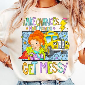May include: A white t-shirt with a colorful graphic featuring a cartoon character with red hair and a yellow school bus. The text on the shirt reads "Take Chances Make Mistakes Get Messy".
