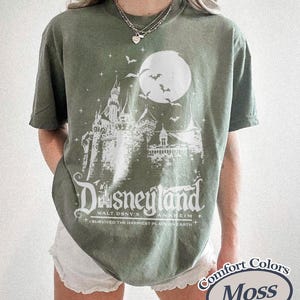 K&ouml;nnte beinhalten: Moosgr&uuml;nes T-Shirt mit wei&szlig;er Grafik eines Schlosses, Mondes und Flederm&auml;usen. Der Text "Disneyland" und "I survived the happiest place on earth" sind darunter gedruckt. Das Shirt ist von der Marke Comfort Colors.