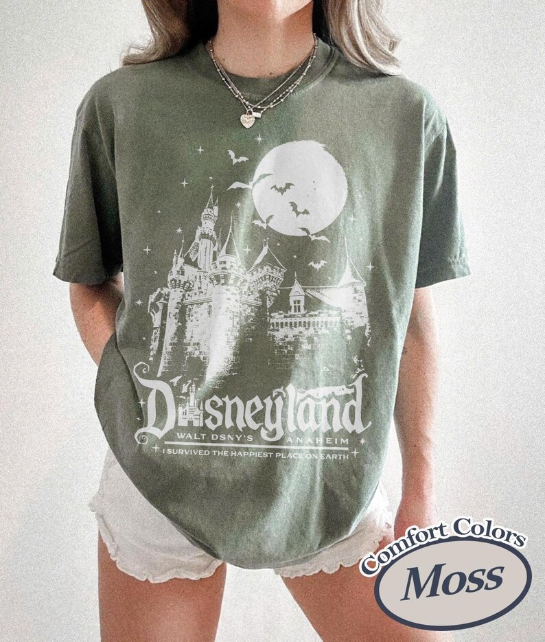 K&ouml;nnte beinhalten: Moosgr&uuml;nes T-Shirt mit wei&szlig;er Grafik eines Schlosses, Mondes und Flederm&auml;usen. Der Text "Disneyland" und "I survived the happiest place on earth" sind darunter gedruckt. Das Shirt ist von der Marke Comfort Colors.