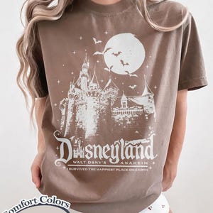 K&ouml;nnte beinhalten: Braunes T-Shirt mit wei&szlig;er Grafik eines Schlosses, Mondes und Flederm&auml;usen, mit dem Wort "Disneyland" und "Walt Dsny's Anaheim". Das T-Shirt enth&auml;lt auch den Satz "I survived the happiest place on earth".