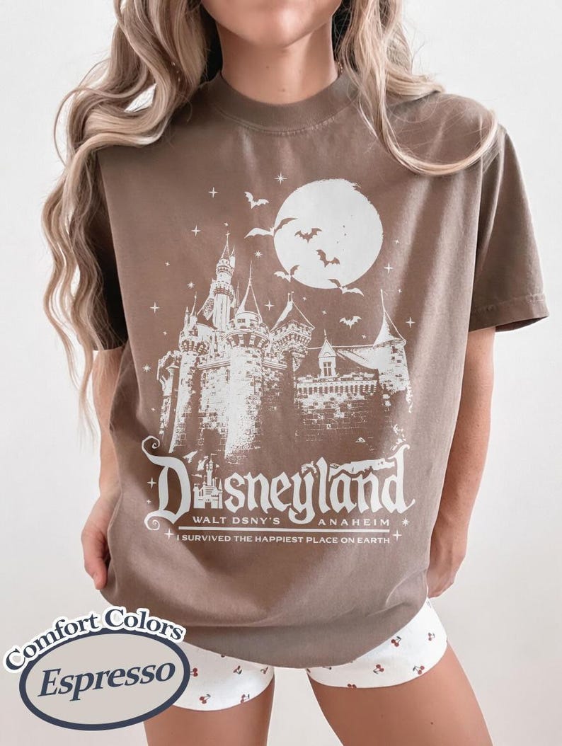 K&ouml;nnte beinhalten: Braunes T-Shirt mit wei&szlig;er Grafik eines Schlosses, Mondes und Flederm&auml;usen, mit dem Wort "Disneyland" und "Walt Dsny's Anaheim". Das T-Shirt enth&auml;lt auch den Satz "I survived the happiest place on earth".