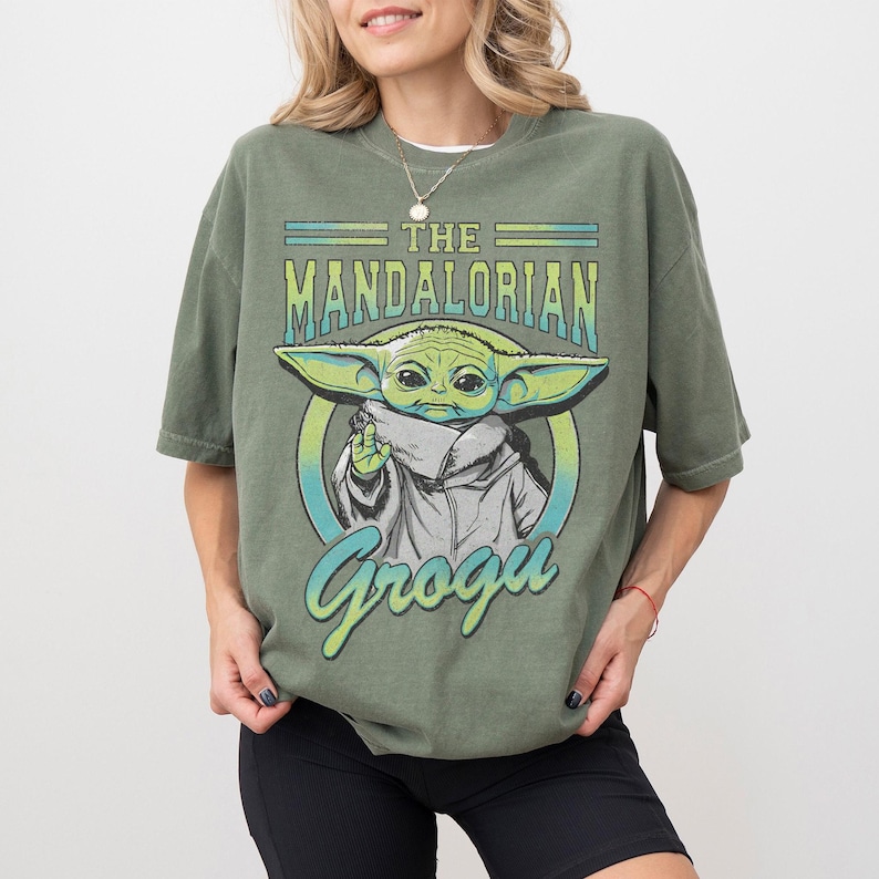 May include: An olive green short-sleeve t-shirt featuring a graphic print of Grogu from The Mandalorian. The design includes the text THE MANDALORIAN in a bold font above the character and the name Grogu in a script font below the character.
