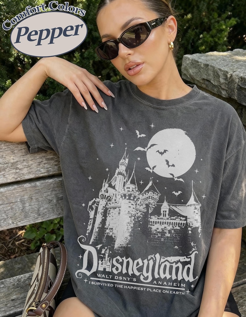 K&ouml;nnte beinhalten: Dunkelgraues T-Shirt mit einem Disneyland-Motiv. Das Design zeigt ein Schloss, Flederm&auml;use und einen Vollmond. Der Text lautet "Disneyland Walt DSNY's Anaheim" und "I survived the happiest place on earth."