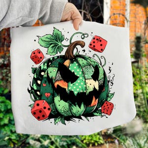 Könnte beinhalten: Weiße Tragetasche mit einem grün-schwarzen Patchwork-Kürbis-Design, das mit einem gruseligen Gesicht geschnitzt ist. Rote Würfel und grüne Ranken umgeben den Kürbis. Ein brauner Stiel und grüne Blätter vervollständigen das Halloween-Thema.