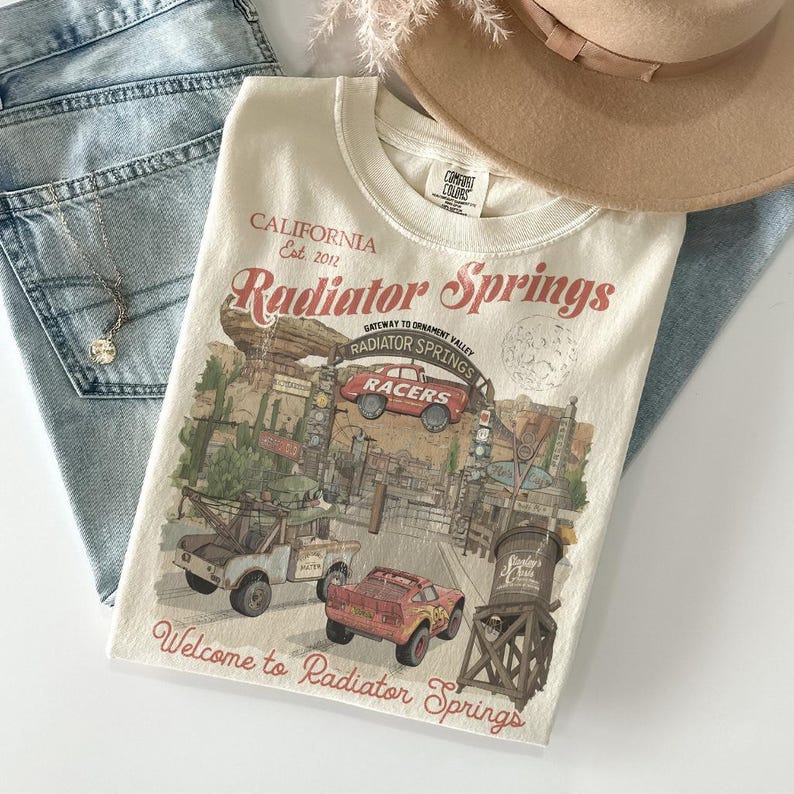 May include: Cream-colored t-shirt featuring a graphic of Radiator Springs, with text that reads "CALIFORNIA Est. 2012 Radiator Springs" and "Welcome to Radiator Springs." The design includes illustrations of cars and buildings. The shirt is paired with light-wash denim jeans and a tan hat.