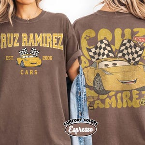 May include: Brown t-shirt featuring a yellow cartoon car with checkered flags, the name "Cruz Ramirez," and the word "Cars." The front of the shirt has the text "EST. 2006." The back of the shirt has a larger graphic of the car and the text "Route 66."
