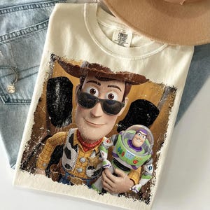 K&ouml;nnte beinhalten: Ein cremefarbenes T-Shirt mit einem Grafikdruck der Figuren Woody und Buzz Lightyear. Woody tr&auml;gt eine schwarze Sonnenbrille und einen braunen Cowboyhut. Der Druck hat ein abgenutztes, strukturiertes Aussehen mit dunklen R&auml;ndern. Das Shirt hat ein Comfort Colors Etikett.