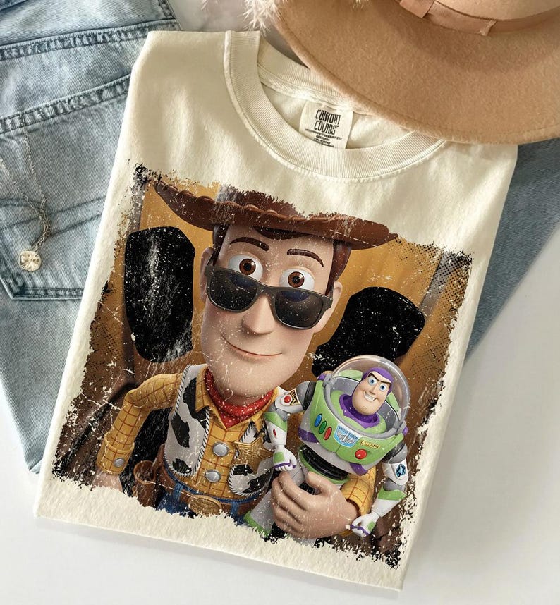K&ouml;nnte beinhalten: Ein cremefarbenes T-Shirt mit einem Grafikdruck der Figuren Woody und Buzz Lightyear. Woody tr&auml;gt eine schwarze Sonnenbrille und einen braunen Cowboyhut. Der Druck hat ein abgenutztes, strukturiertes Aussehen mit dunklen R&auml;ndern. Das Shirt hat ein Comfort Colors Etikett.