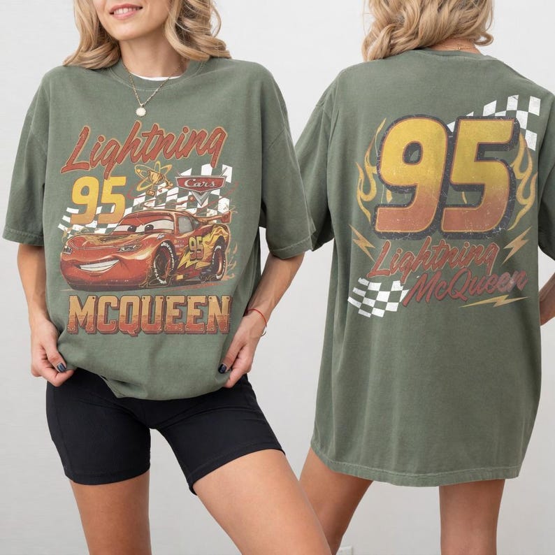 Op de afbeelding: Een olijfgroen T-shirt met korte mouwen en een afbeelding van de rode raceauto Lightning McQueen op de voorkant met de tekst Lightning 95 MCQUEEN. De achterkant toont het nummer 95 in een groot geel lettertype met vlammen en een geblokt patroon.