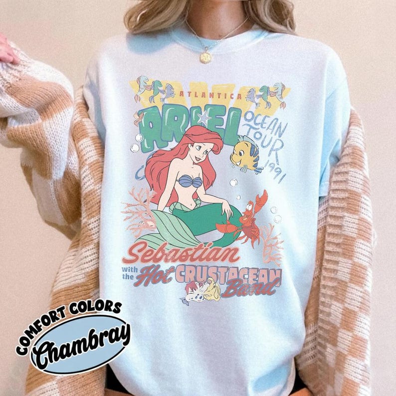 May include: A light blue crewneck sweatshirt featuring a graphic print of Ariel, Flounder, and Sebastian from The Little Mermaid. The text reads Atlantica Ariel Ocean Tour 1991, Sebastian with the Hot Crustacean Band. A beige and white checkered cardigan is layered over it.