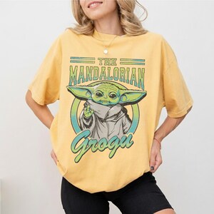 May include: A yellow short-sleeve t-shirt featuring a graphic print of Grogu from The Mandalorian. The design includes the text THE MANDALORIAN and Grogu in a teal and green color palette, centered on the chest of the garment with a circular frame.