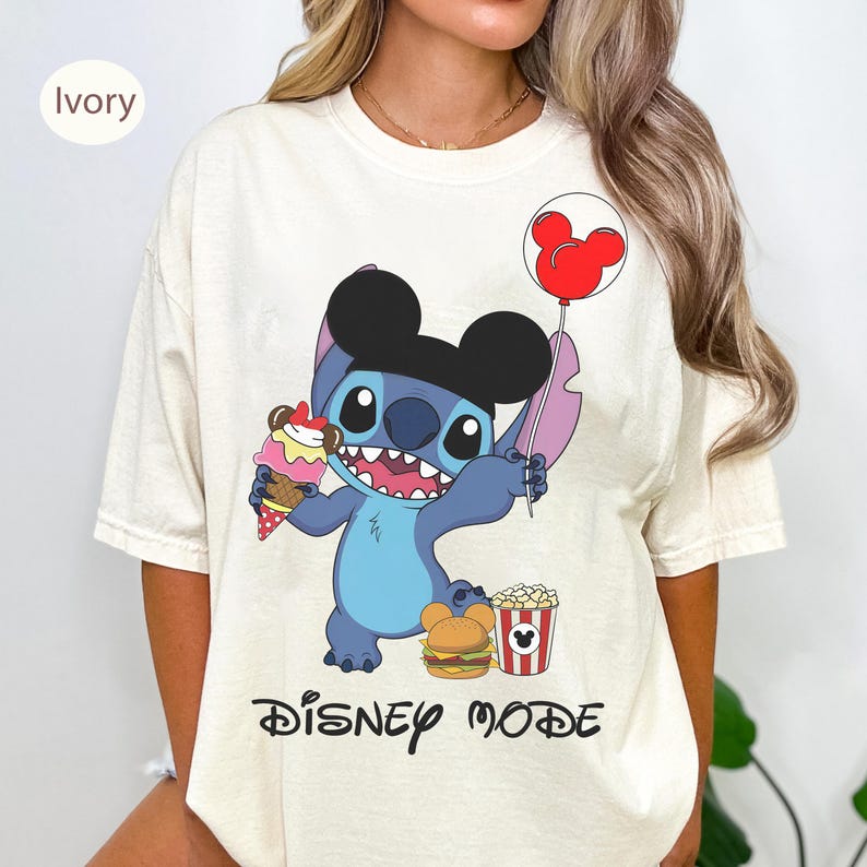 Puede incluir: Camiseta color marfil con un personaje de dibujos animados sosteniendo un helado y un globo de Mickey Mouse. El personaje azul con orejas negras est&aacute; rodeado de una hamburguesa y palomitas de ma&iacute;z. Las palabras "DISNEY MODE" est&aacute;n impresas debajo.