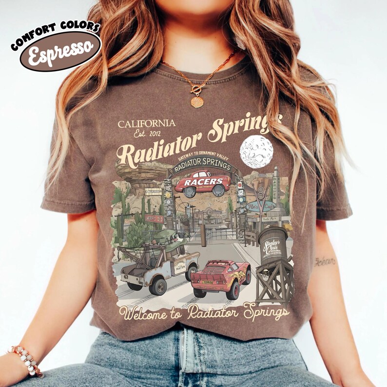 May include: Espresso-colored t-shirt featuring a graphic of Radiator Springs, with the text "Welcome to Radiator Springs." The design includes illustrations of cars, buildings, and the text "California Est. 2012."