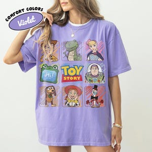 May include: A lilac Comfort Colors t-shirt with a Toy Story character grid design. The shirt features Woody, Rex, Bo Peep, Buzz Lightyear, Slinky Dog, Jessie, and Mr. Pricklepants. The words "Hi!" and "Toy Story" are also visible.