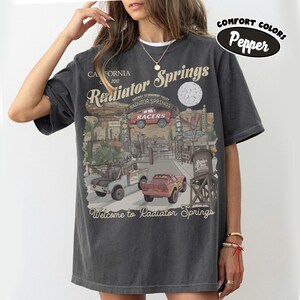 May include: Dark grey t-shirt featuring a graphic of Radiator Springs, with the text "Radiator Springs" and "Welcome to Radiator Springs". The design includes images of cars and a desert landscape. The shirt is labeled "Comfort Colors Pepper".