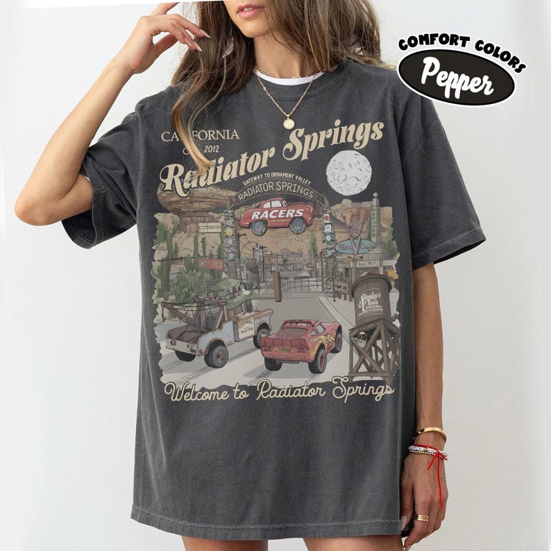 May include: Dark grey t-shirt featuring a graphic of Radiator Springs, with the text "Radiator Springs" and "Welcome to Radiator Springs". The design includes images of cars and a desert landscape. The shirt is labeled "Comfort Colors Pepper".