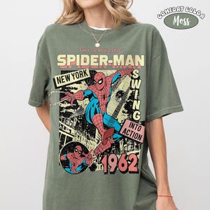 Puede incluir: Una camiseta de manga corta color verde musgo con un estampado gr&aacute;fico de Spider-Man en rojo y azul. El texto dice THE AMAZING SPIDER-MAN, NEW YORK, SWING INTO ACTION y 1962. Una peque&ntilde;a insignia circular en la esquina superior derecha dice COMFORT COLOR MOSS.