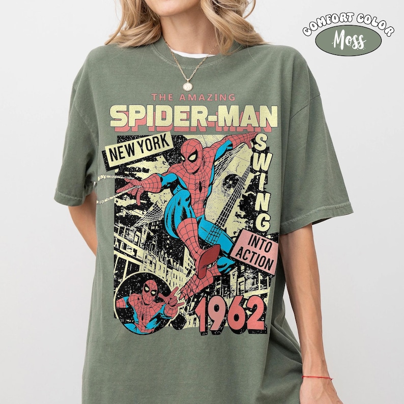 Puede incluir: Una camiseta de manga corta color verde musgo con un estampado gr&aacute;fico de Spider-Man en rojo y azul. El texto dice THE AMAZING SPIDER-MAN, NEW YORK, SWING INTO ACTION y 1962. Una peque&ntilde;a insignia circular en la esquina superior derecha dice COMFORT COLOR MOSS.