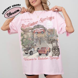 May include: A light pink t-shirt with a graphic of Radiator Springs, featuring cars and the text "Welcome to Radiator Springs." The shirt also has the text "California Est. 2012" and "Comfort Colors Blossom."