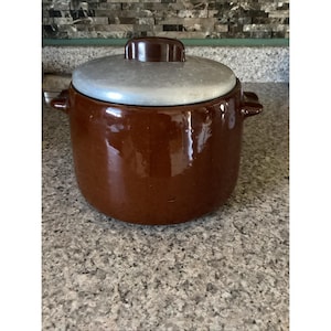 May include: A brown ceramic pot with a lid. The pot has two handles and a shiny, glazed finish. The lid is a light gray color with a brown knob. The pot is sitting on a speckled countertop.