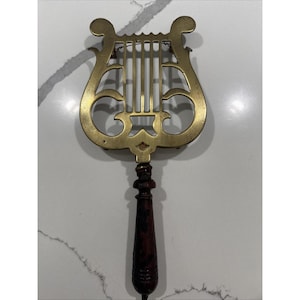 May include: A vintage brass music stand in the shape of a lyre, featuring a dark wooden handle. The brass has a warm, golden hue, and the handle is a rich, dark brown. The stand is designed to hold sheet music.