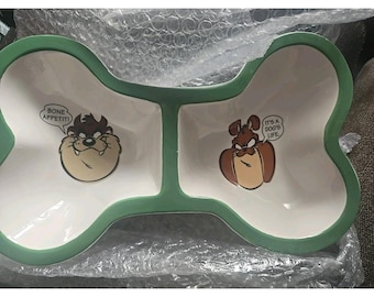 Puede incluir: Cuenco para mascotas con forma de hueso y dos compartimentos. El cuenco es blanco con un borde verde. Un lado presenta a Taz de Looney Tunes con el texto "BONE APPETIT!" y el otro lado un perro de dibujos animados con el texto "IT'S A DOG'S LIFE."