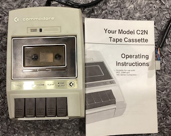 Può includere: Un lettore di cassette Commodore C2N beige con cavo di alimentazione nero e cinque pulsanti di controllo marrone scuro, accompagnato da un manuale bianco intitolato Your Model C2N Tape Cassette Operating Instructions per computer.
