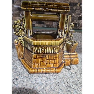 Può includere: Una fioriera a forma di pozzo dei desideri in ceramica nei toni del marrone e del verde. Il pozzo presenta un design a forma di mattone, un tetto e un piccolo secchio appeso a una catena. La parola "WISHING" è incisa sulla parte anteriore.