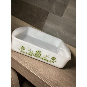 Könnte beinhalten: Eine rechteckige Backform aus weißem Milchglas mit einem grünen Blumenmuster auf der Vorderseite. Die Form verfügt über integrierte Griffe an den kurzen Seiten und eine glatte, glänzende Oberfläche. Das Design besteht aus stilisierten grünen Blumen und Ranken.