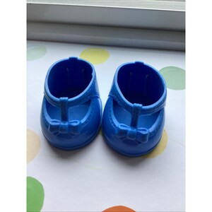 May include: A pair of bright blue plastic doll shoes featuring a rounded toe, a T-strap design, and a small molded bow detail at the center of the strap. The shoes have a smooth, glossy finish and are designed with an open top for easy access.