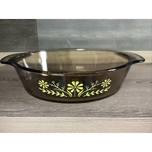 Pode incluir: Uma assadeira redonda de vidro castanho translúcido com pegas laterais integradas. O exterior apresenta um padrão repetitivo de flores amarelo-brilhante e caules de folhas delicadas impressos horizontalmente no centro da taça.