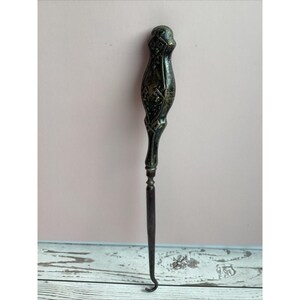 May include: An antique metal crochet hook with an ornate handle. The handle is dark with engraved floral and geometric patterns. The hook is a dark metal color and has a curved end. The hook is displayed against a pink and white background.