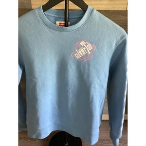 May include: Light blue Levi's sweatshirt with a crew neck. The front features a pink and white graphic of a globe with the text "silver lab". The sleeve has white text. The sweatshirt is hanging on a black hanger.