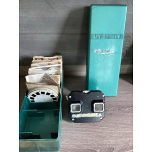 May include: A vintage View-Master set, including a black viewer, a teal storage case with "View-Master" text, and a teal storage box filled with image reels. The set is on a wooden surface.