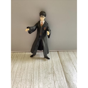 May include: A plastic figurine of a character from the Harry Potter series. The figure is wearing a black robe, gray pants, and black shoes. The figure has black hair, glasses, and a tie. The figure is standing on a wooden surface.